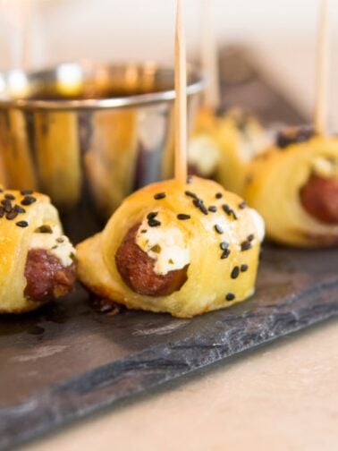 Little smoky hot dogs wrapped in dough and sprinkled with sesame seeds all on slate serving platter on marble surface
