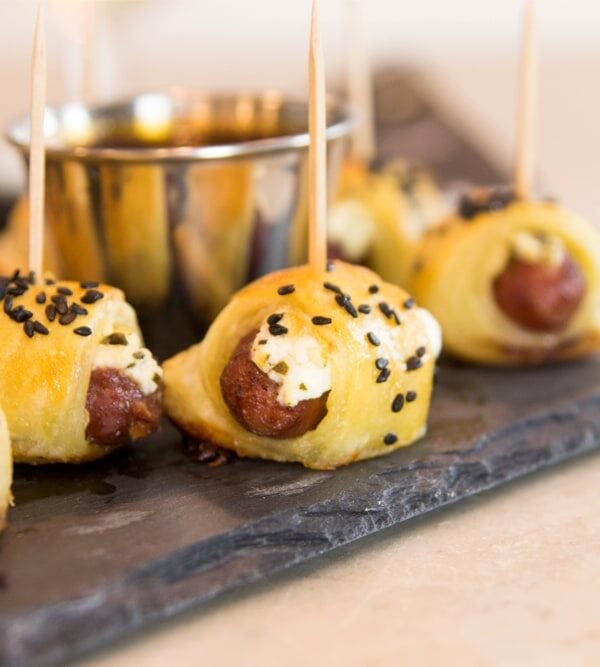 Little smoky hot dogs wrapped in dough and sprinkled with sesame seeds all on slate serving platter on marble surface