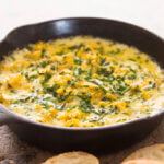 Cast-iron skillet filled with butternut squash cheese dip with herbs on top sitting on wooden board with a few slices of toasted bread showing