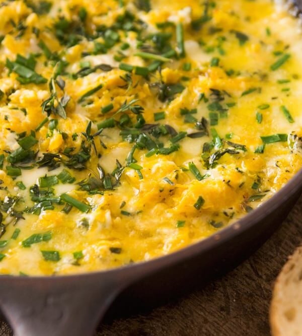 Cast-iron skillet filled with light yellow colored butternut squash cheese dip with sprinkling of herbs on top with slices of bread on one side