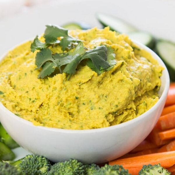 White bowl filled with yellow-colored curried white bean hummus with cilantro leaves on top surrounded by sliced vegetables