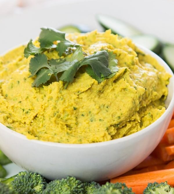 White bowl filled with yellow-colored curried white bean hummus with cilantro leaves on top surrounded by sliced vegetables