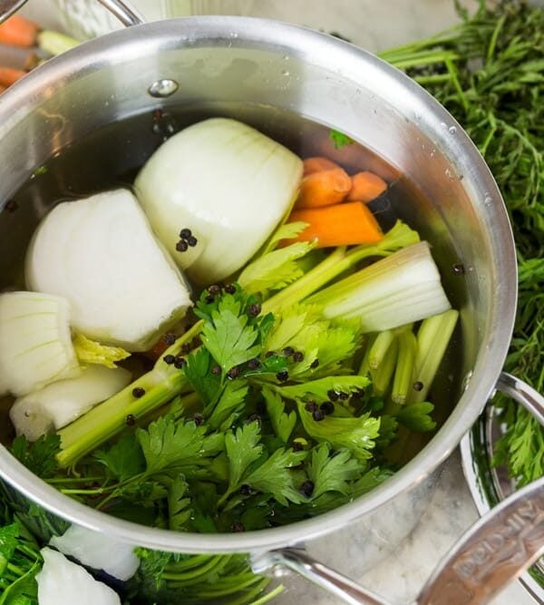 Large steel pot sitting on marble surface filled with greens, onions, carrots, and peppercorns along with lots of water.