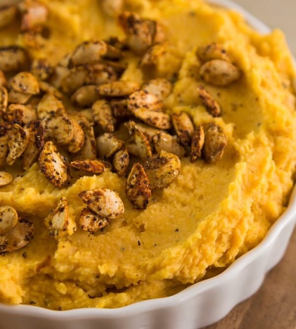 White container filled with butternut squash hummus topped with spice-covered pepitas sitting on white marble surface