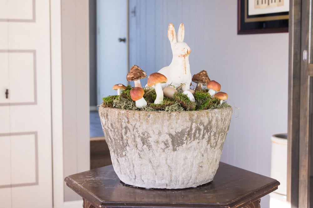 Concrete large vessel sitting on a wooden table decorated with cast iron bunny and small mushrooms for an Easter display.