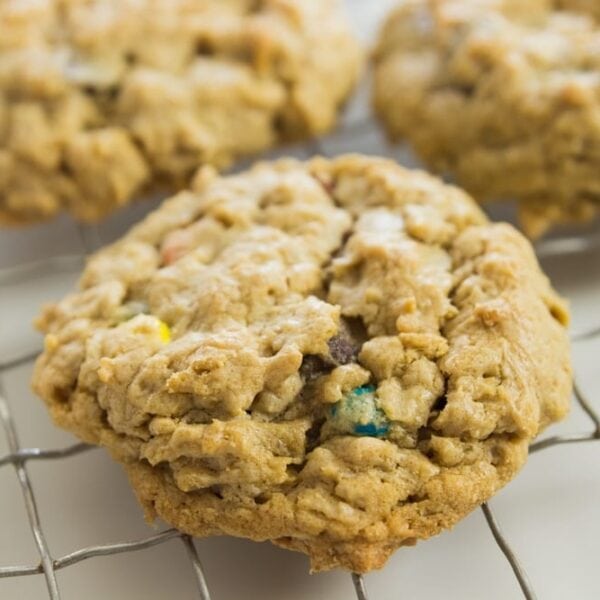 Large monster cookie made from oats and M&M candies sitting on wire cooling rack on white surface with two other cookies in background