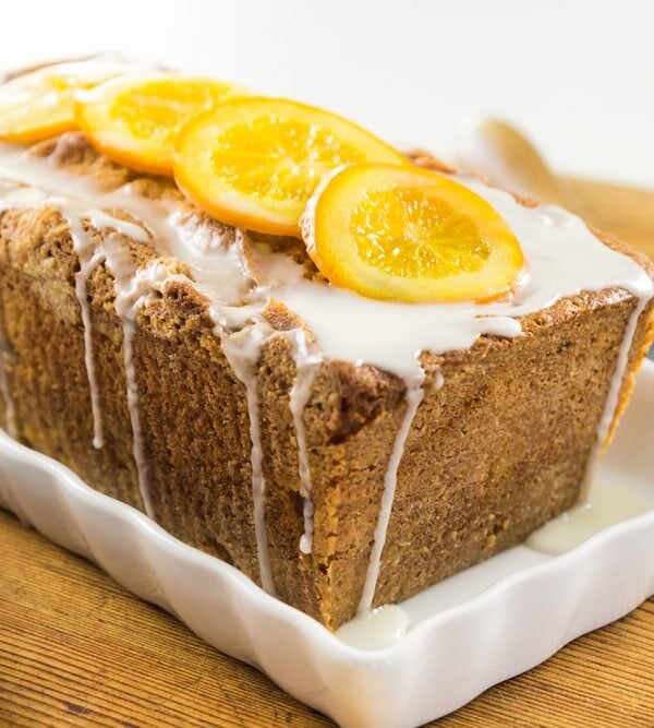 Pound cake loaf sitting on white serving platter on wood board with slicing knife behind topped with orange slices and a white glaze