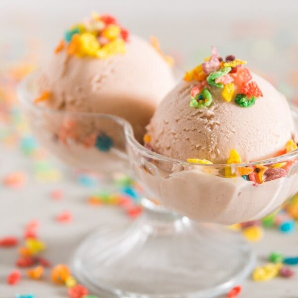 Fruity Pebbles cereal sprinkled on top of pink ice cream in a double-scoop bowl with Fruity Pebbles cereal in the background