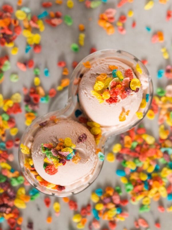 Top down view of Fruity Pebbles cereal sprinkled on top of pink ice cream in a double-scoop bowl with Fruity Pebbles cereal in the background
