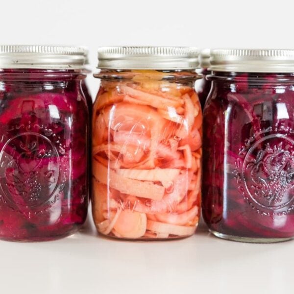 Three different kinds of canned beet recipes in Ball canning jars on a white surface with red and yellow beets in the background.