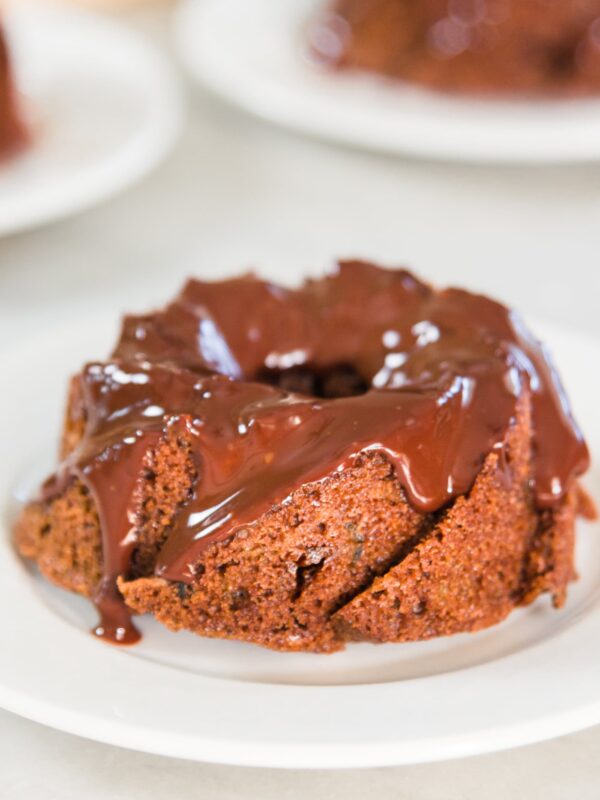 Mini brown Bundt chocolate cake dripping with icing sitting on white plate on marble surface with other small cakes in background