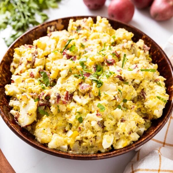 Brown bowl sitting on white surface filled with yellow potato salad topped with parsley with red potatoes and parsley with wooden spoon beside.