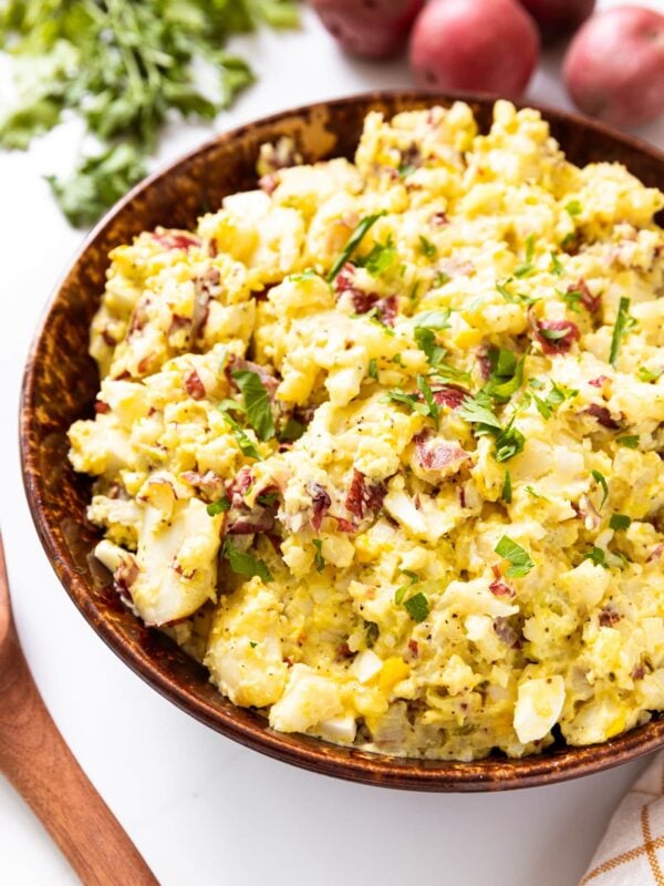 Brown bowl filled with yellow colored potato salad topped with a sprinkling of parsley with red potatoes and parsley in background all on white surface.
