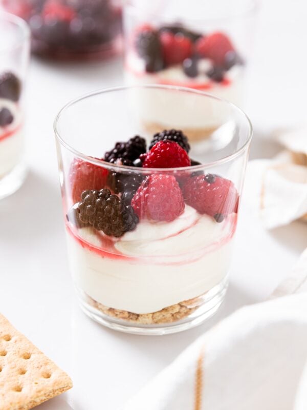 Glass container filled with triple berry cheesecake with graham cracker base all sitting on white countertop with more glasses in background.