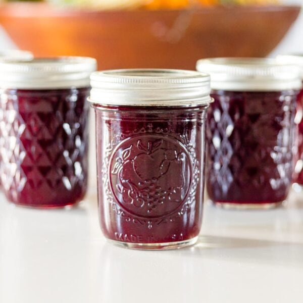 Five jam jars full of can grape butter with a brown bowl in background on a white surface