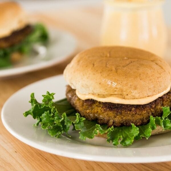 Quinoa squash burger sitting on white plate with spicy aioli sauce in between bun and lettuce all on wood surface with second burger in background