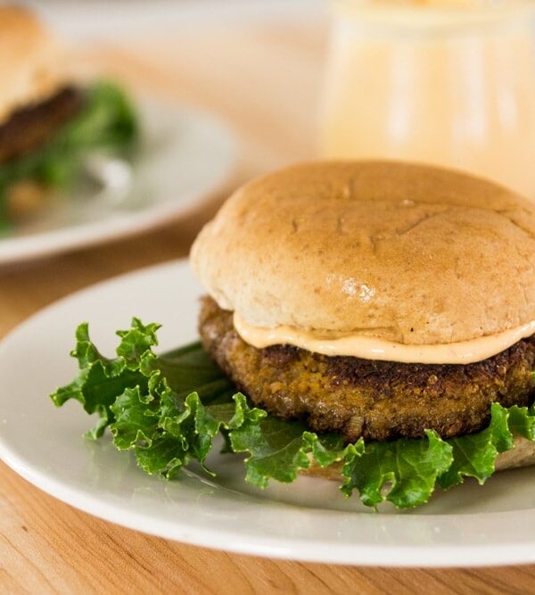 Quinoa squash burger sitting on white plate with spicy aioli sauce in between bun and lettuce all on wood surface with second burger in background