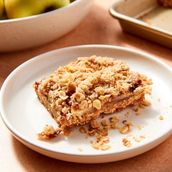 White plate with slice of apple pie bar with rest of bar in the background along with a bowl of apples.