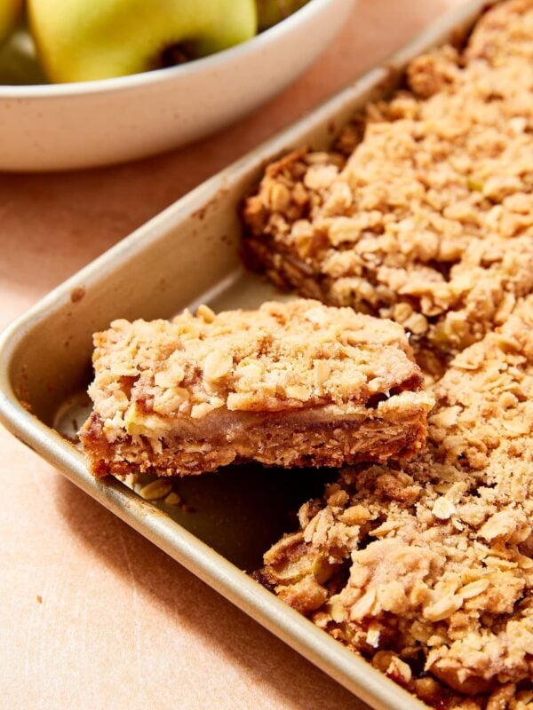Gold pan filled with baked apple pie bars with streusel topping with bowl of apples in the background.