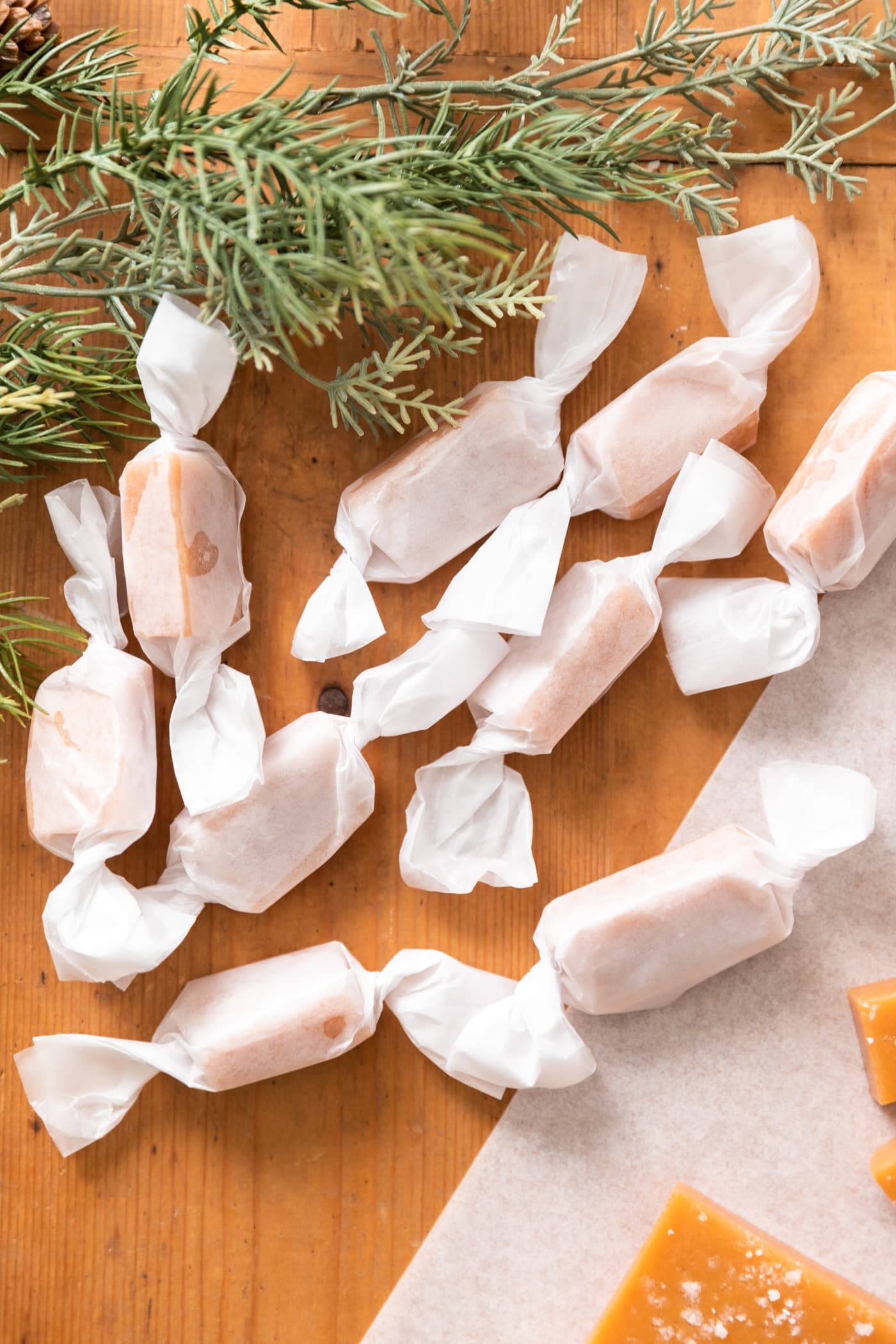 Wax wrapped pieces of caramel sitting on wood surface with greenery in background and unwrapped caramels beside