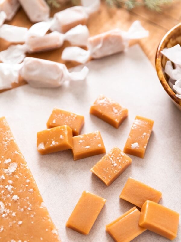 Pieces of golden cut caramel sitting on white piece of parchment paper with wrapped caramels in wax paper in background