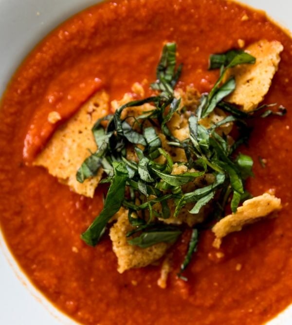 Top down view of white bowl filled with red colored tomato and roasted red pepper soup with Parmesan crisp and strips on basil on top