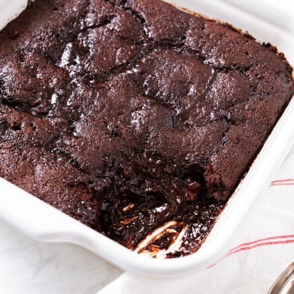 Square white baking dish filled with baked chocolate fudge pudding cake with one scoop mixing showing the interior texture of the bottom of the cake.