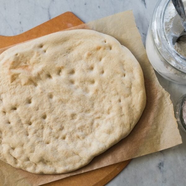 Top down view of baked pizza dough with no toppings sitting on piece of parchment with flour canister all on marble surface