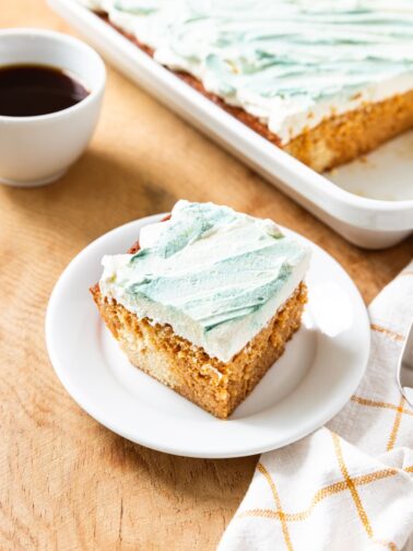 White plate with slice of tres leches cake with whipped topping on top swirled with green coloring with cup of black coffee and rest of cake in the background.