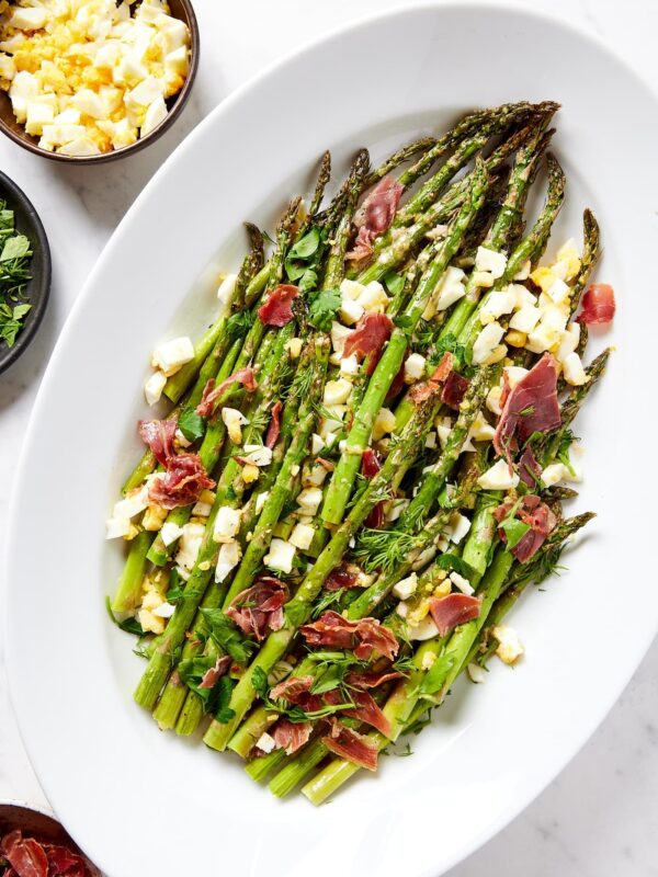 White serving dish filled with roasted asparagus with a lemon vinaigrette drizzled over the top with cooked prosciutto and hardboiled eggs.
