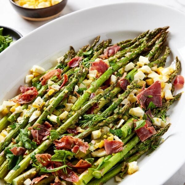White platter filled with pieces of roasted asparagus with chopped hardboiled eggs and pieces of prosciutto.