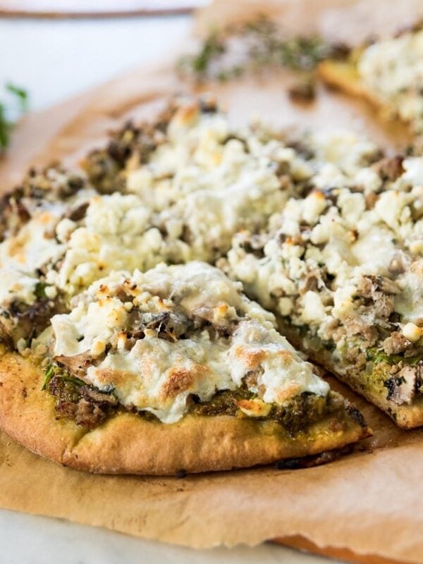 Homemade pizza crust topped with sausage, spinach, and crumbly feta cheese sitting on brown piece of parchment paper on marble surface after coming out of the oven