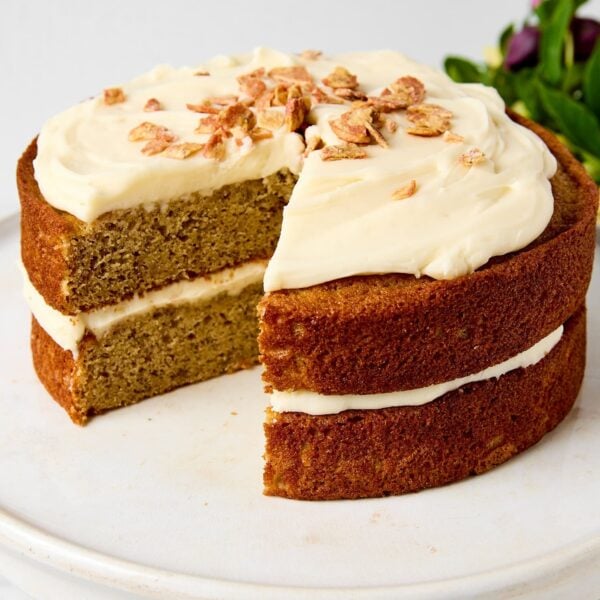 White cake plate with banana cake on top with mascarpone frosting and crushed banana chips.