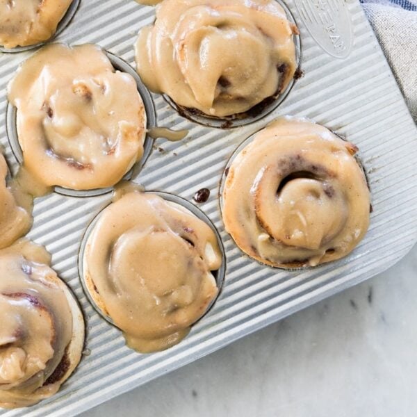 Top down view of silver muffin in containing frosted cinnamon roll muffins with towel on top of marble surface