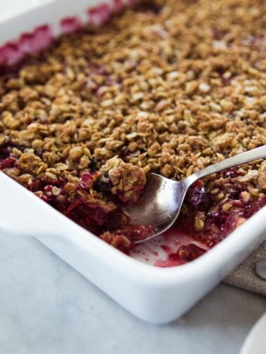 Spoon sitting in white baking dish filled with red colored rhubarb and berry crisp with baked oats on top