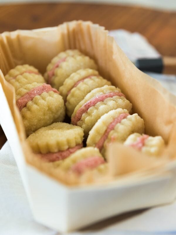 Rhubarb wafer cookies with light pink filling sitting in parchment lined white rectangular box on top of white towel on wood bread board