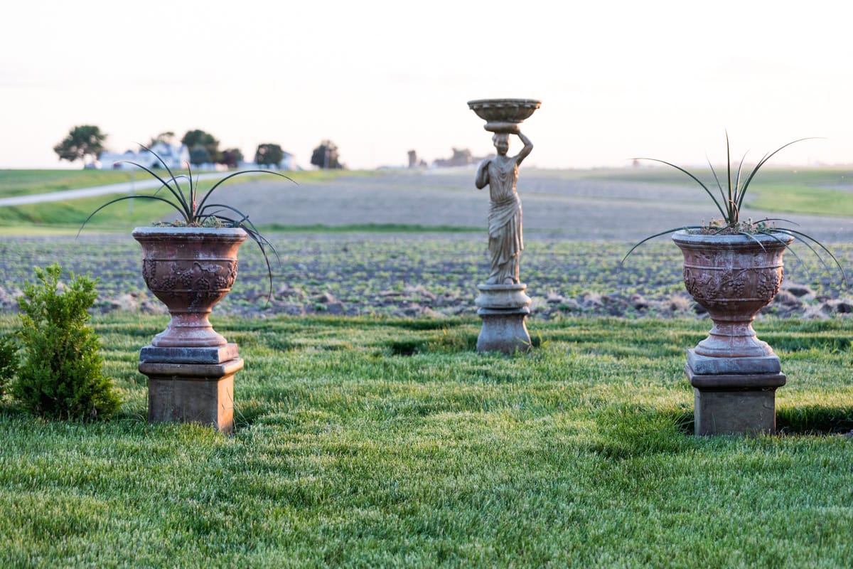 Two urns spaced apart planted with agave with a statue in between all in a yard with fields beyond.