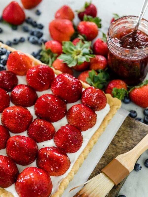 Strawberries and blueberries on a cookie tart with yogurt filling with extra strawberries, blueberries and jam in background on a wood cutting board on a marble surface