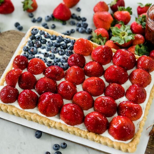 Strawberries and blueberries on a cookie tart with yogurt filling with extra strawberries, blueberries and jam in background on a wood cutting board on a marble surface