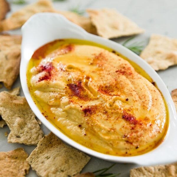 Oval shaped bowl filled with orange-colored traditional hummus topped with paprika with herb crackers all around on marble surface