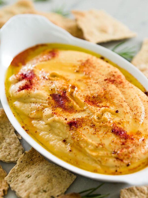 Oval shaped bowl filled with orange-colored traditional hummus topped with paprika with herb crackers all around on marble surface