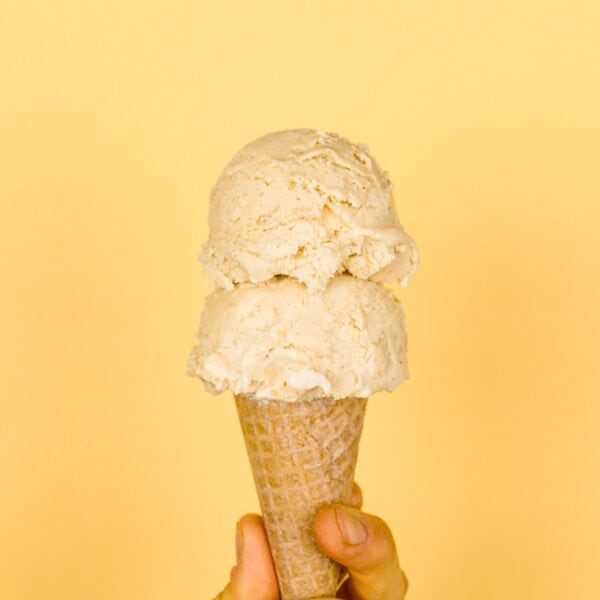 Two scoops of banana cream pie ice cream in ice cream cone held by a hand with light peach colored background