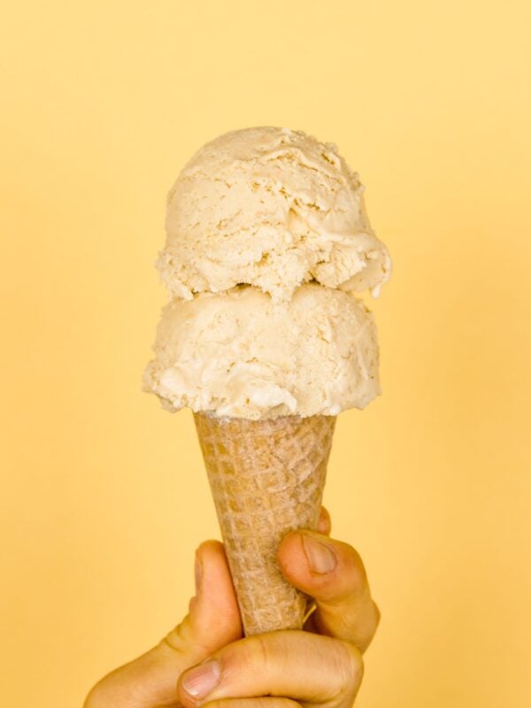 Two scoops of banana cream pie ice cream in ice cream cone held by a hand with light peach colored background