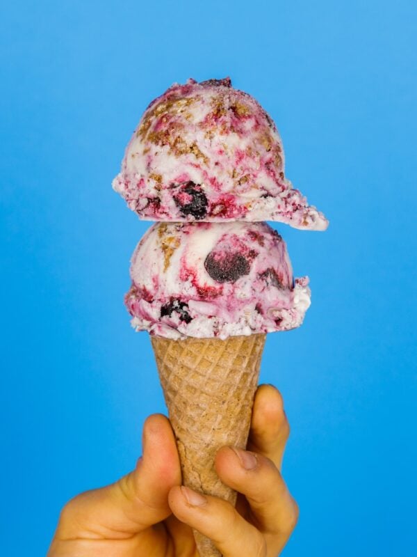 Two scoops of blueberry crumble pie ice cream with swirls of blueberry and homemade streusel on top of a cone being held by a hand with a bright blue background