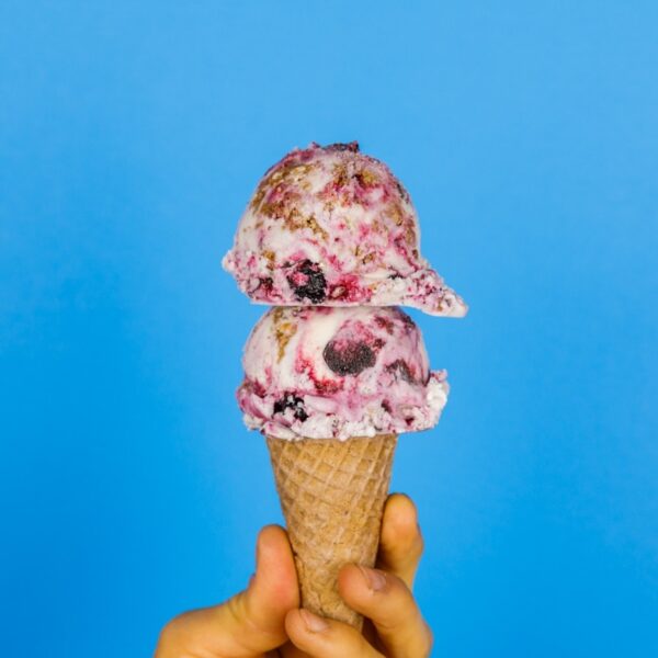 Two scoops of blueberry crumble pie ice cream with swirls of blueberry and homemade streusel on top of a cone being held by a hand with a bright blue background