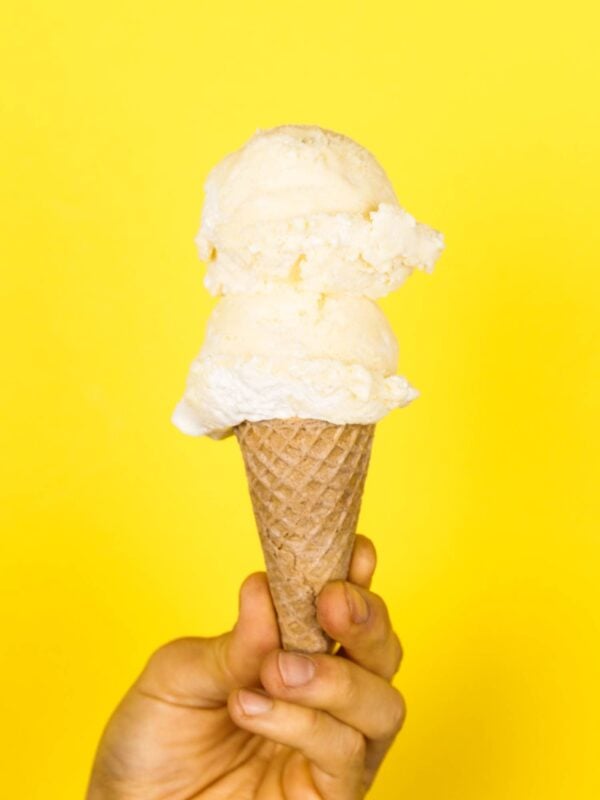 Two scoops of lemon meringue pie ice cream on a cone being held in front of a bright yellow background