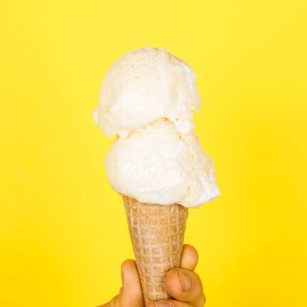 Two scoops of lemon meringue pie ice cream on a cone being held in front of a bright yellow background