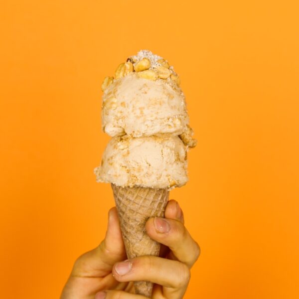 Two scoop of peanut butter pie ice cream on an ice cream cone being held by hand with peanuts and fillings on top in front of orange background