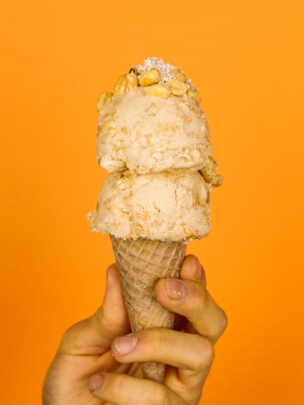 Two scoop of peanut butter pie ice cream on an ice cream cone being held by hand with peanuts and fillings on top in front of orange background