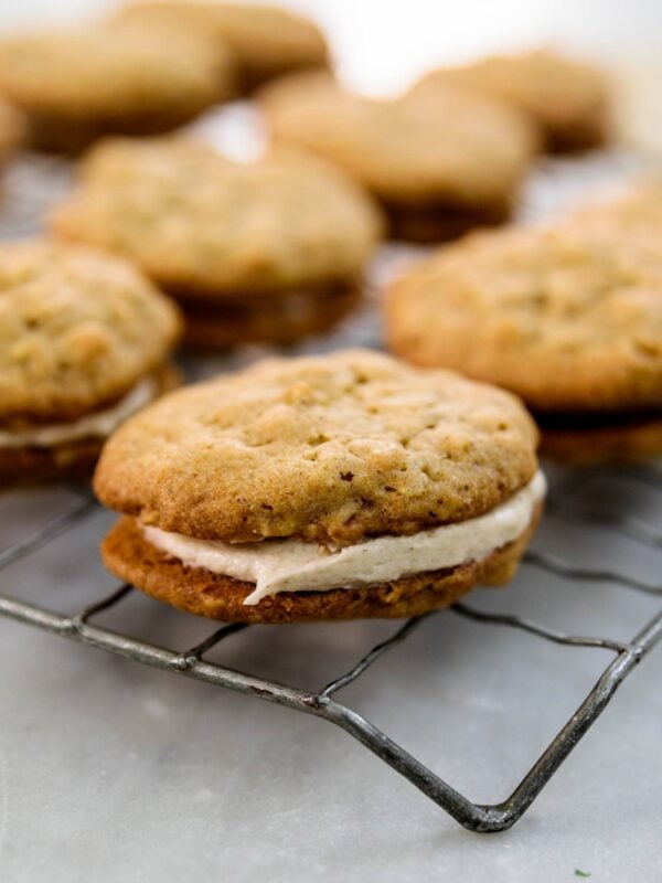 Two oatmeal spice cookies sandwiching a layer of white creme filling with multiple cookies in background all on a wire cooling rack on a marble surface